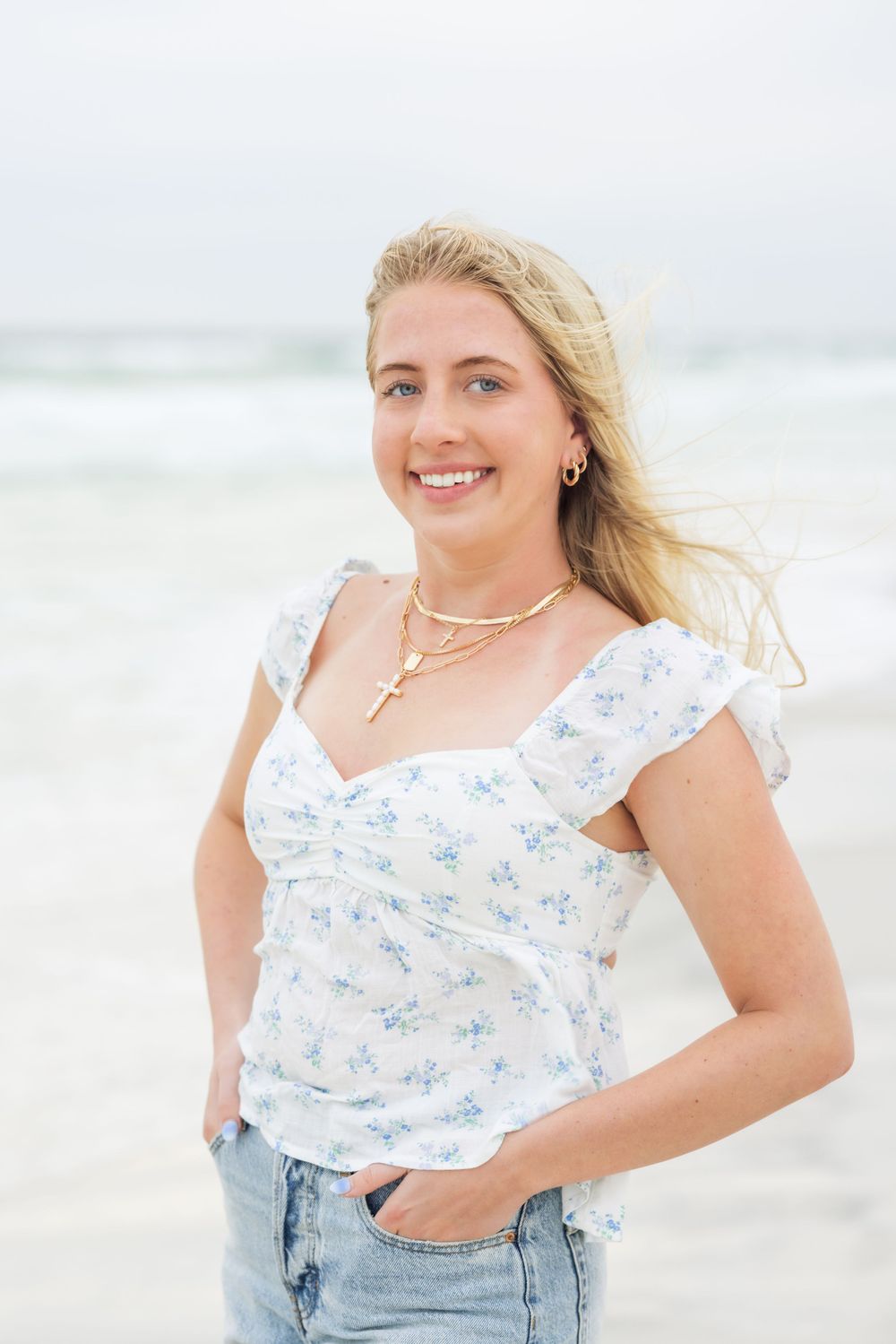 A casual beachside portrait showing someone in a floral top and denim shorts against the ocean backdrop.