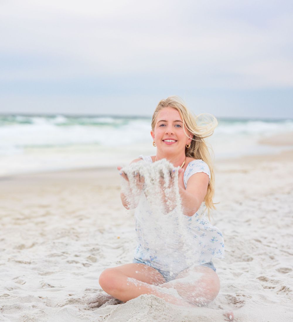 Happy person on a beach tossing white sand into the air while wearing casual summer clothing on a sunny day.