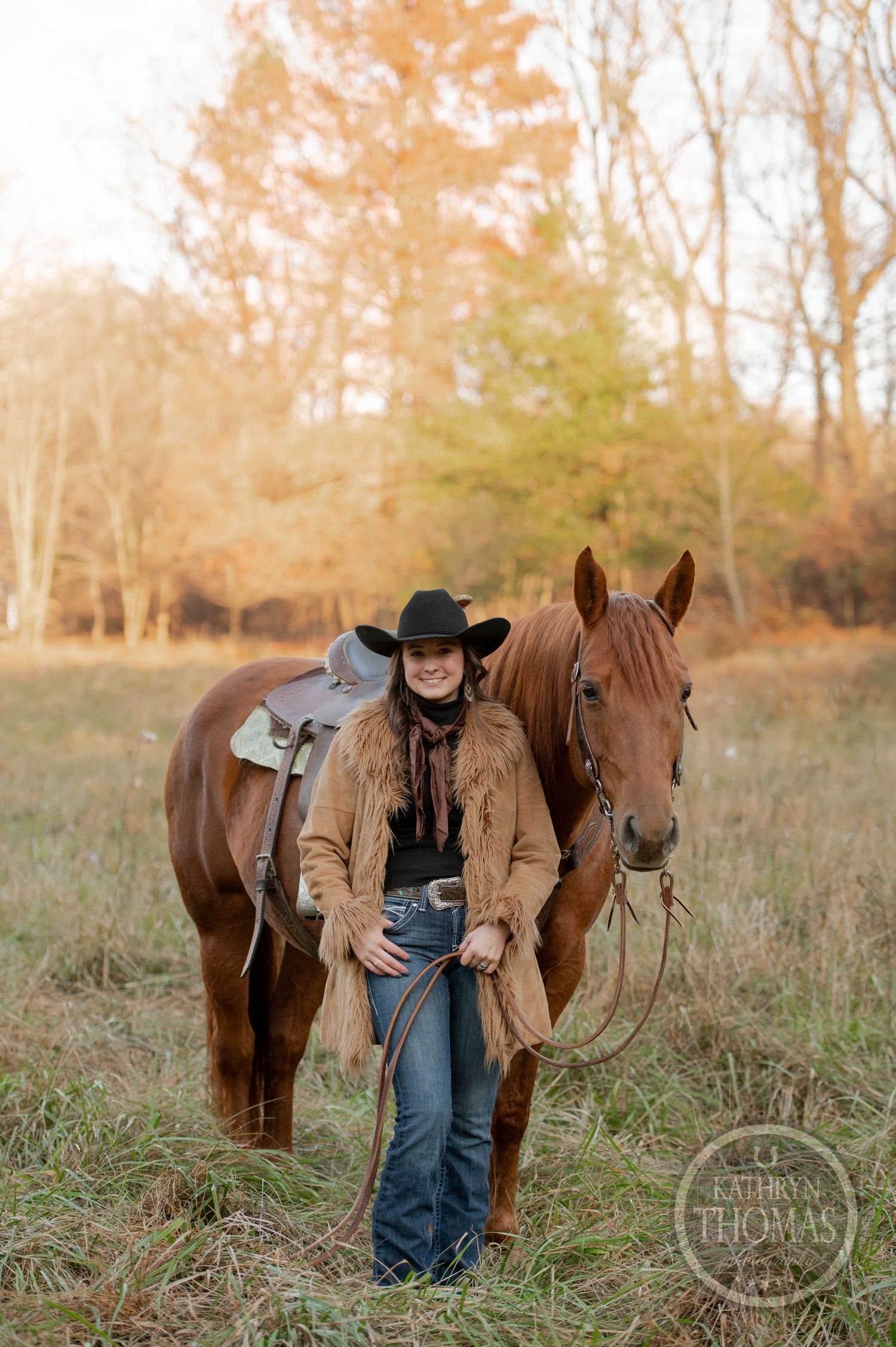 A brown horse wearing a western saddle stands with a rider in a tan leather jacket and black cowboy hat.