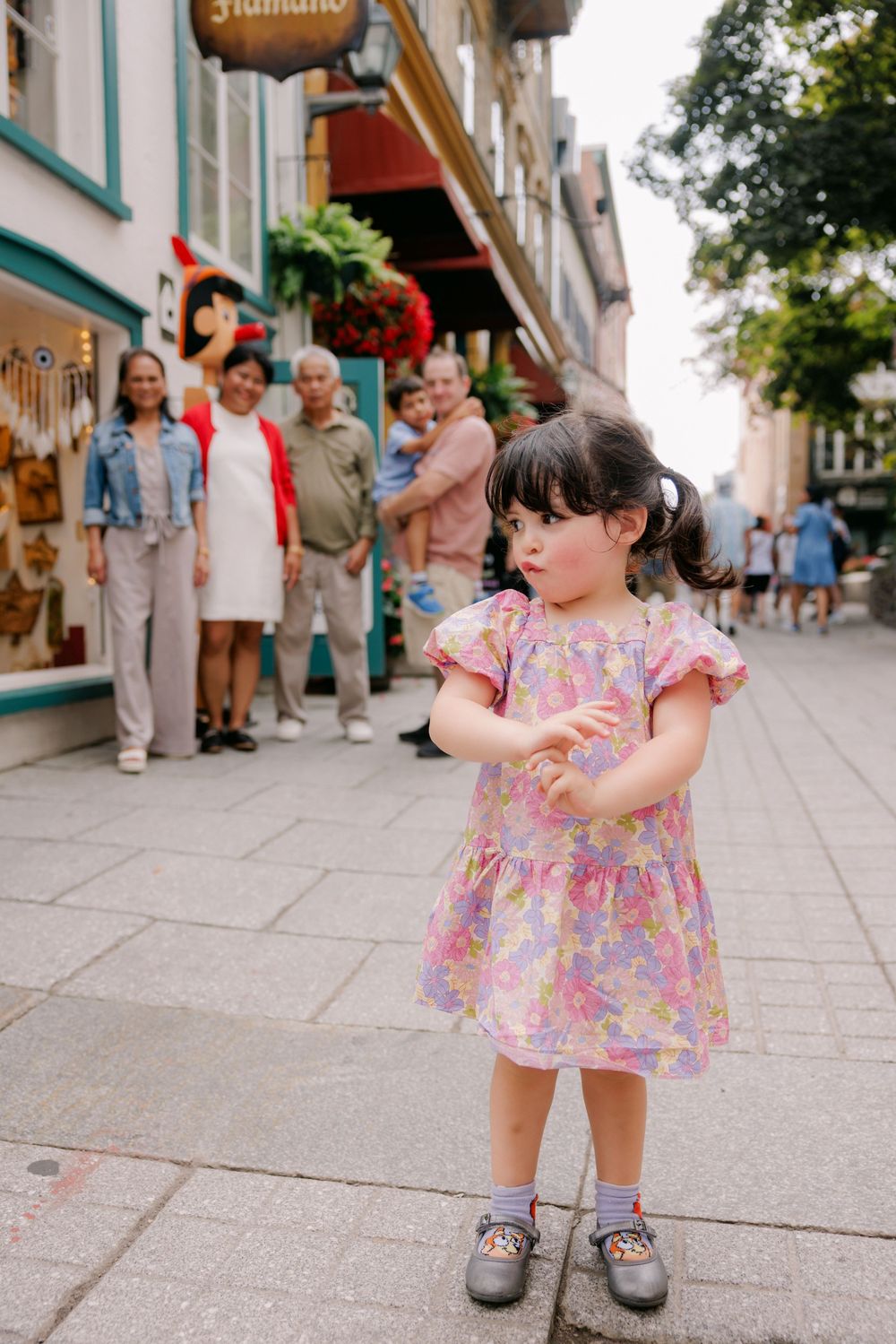 Toddler girl in pink floral dress standing on a cobblestone street with people and colorful shops in the background.
