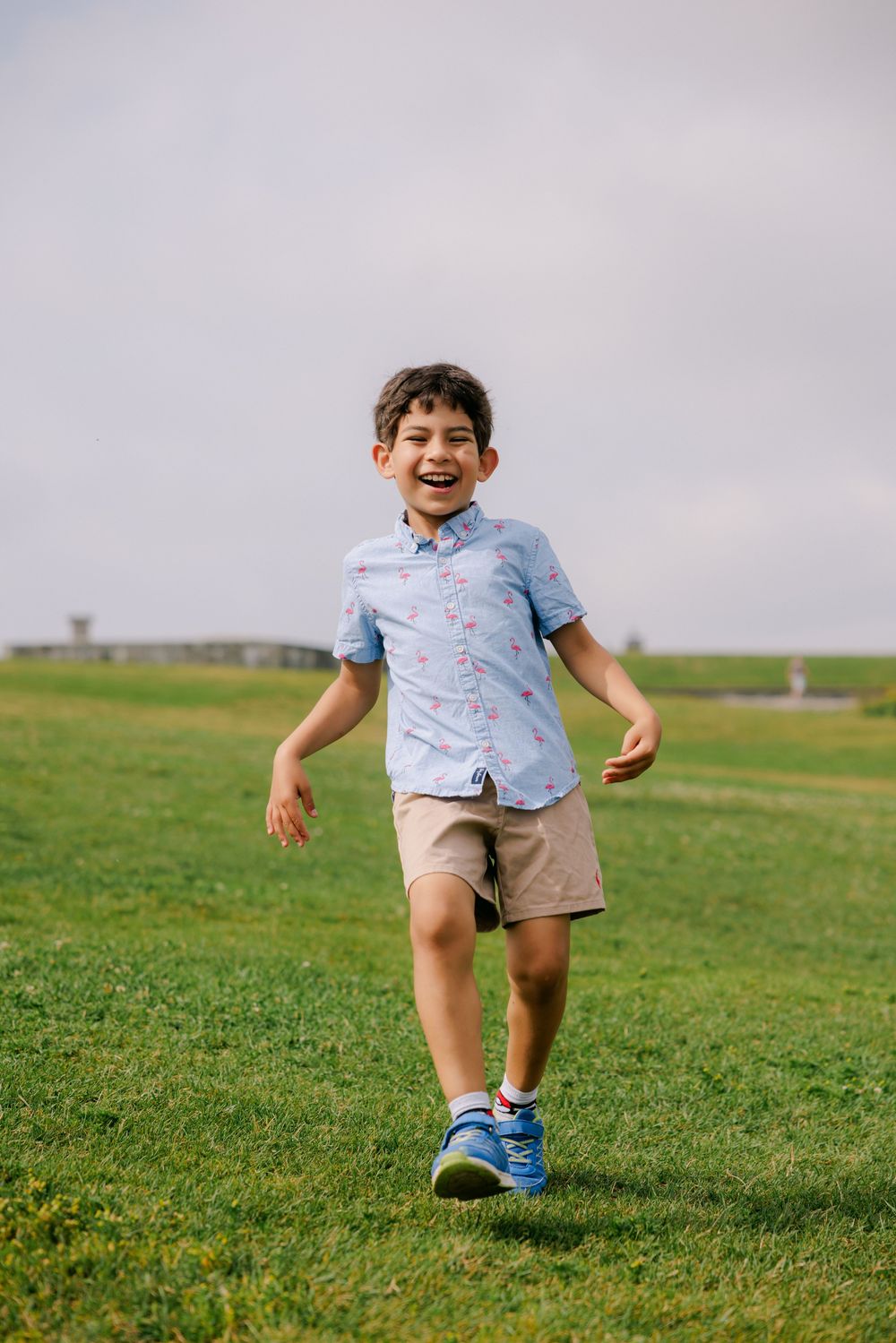 A happy young boy runs across a green grass field wearing a light blue shirt, khaki shorts, and blue sneakers.