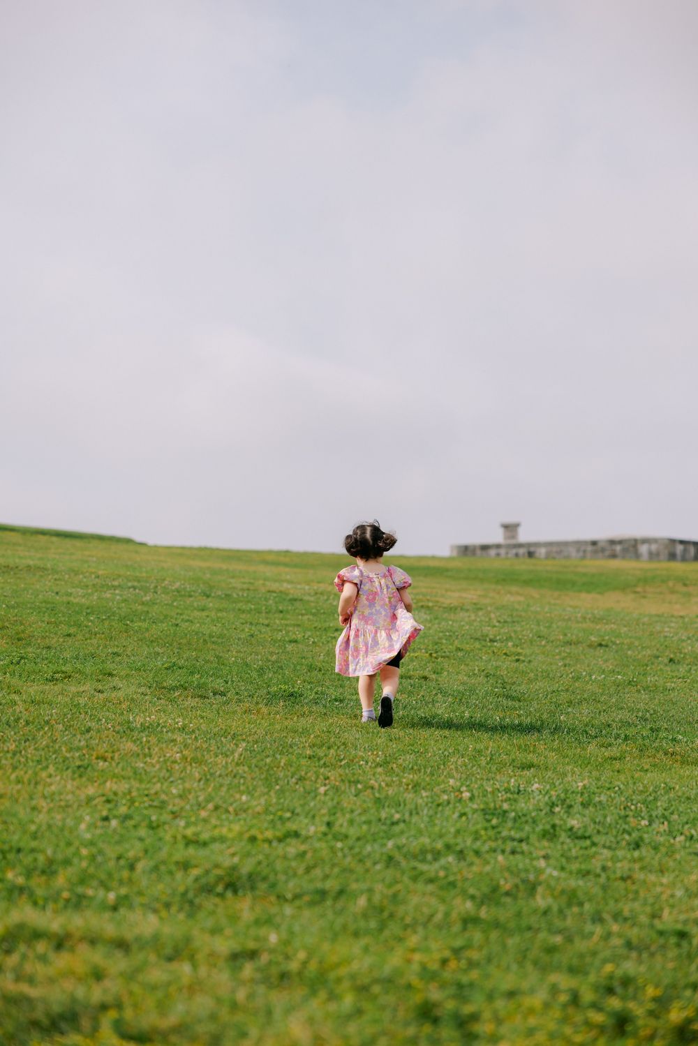 A young girl in a pink dress runs across a lush green field under a cloudy sky.