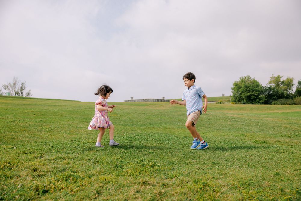 Two children playing and running on a lush green grass field under a cloudy sky on a warm day.