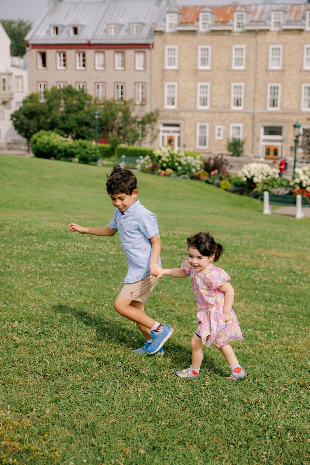 Two young children holding hands and running on green grass in front of a large stone building on a sunny day.