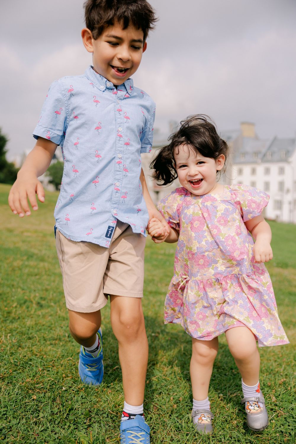 Two happy children holding hands and running on green grass, boy in blue flamingo shirt and girl in pink floral dress.