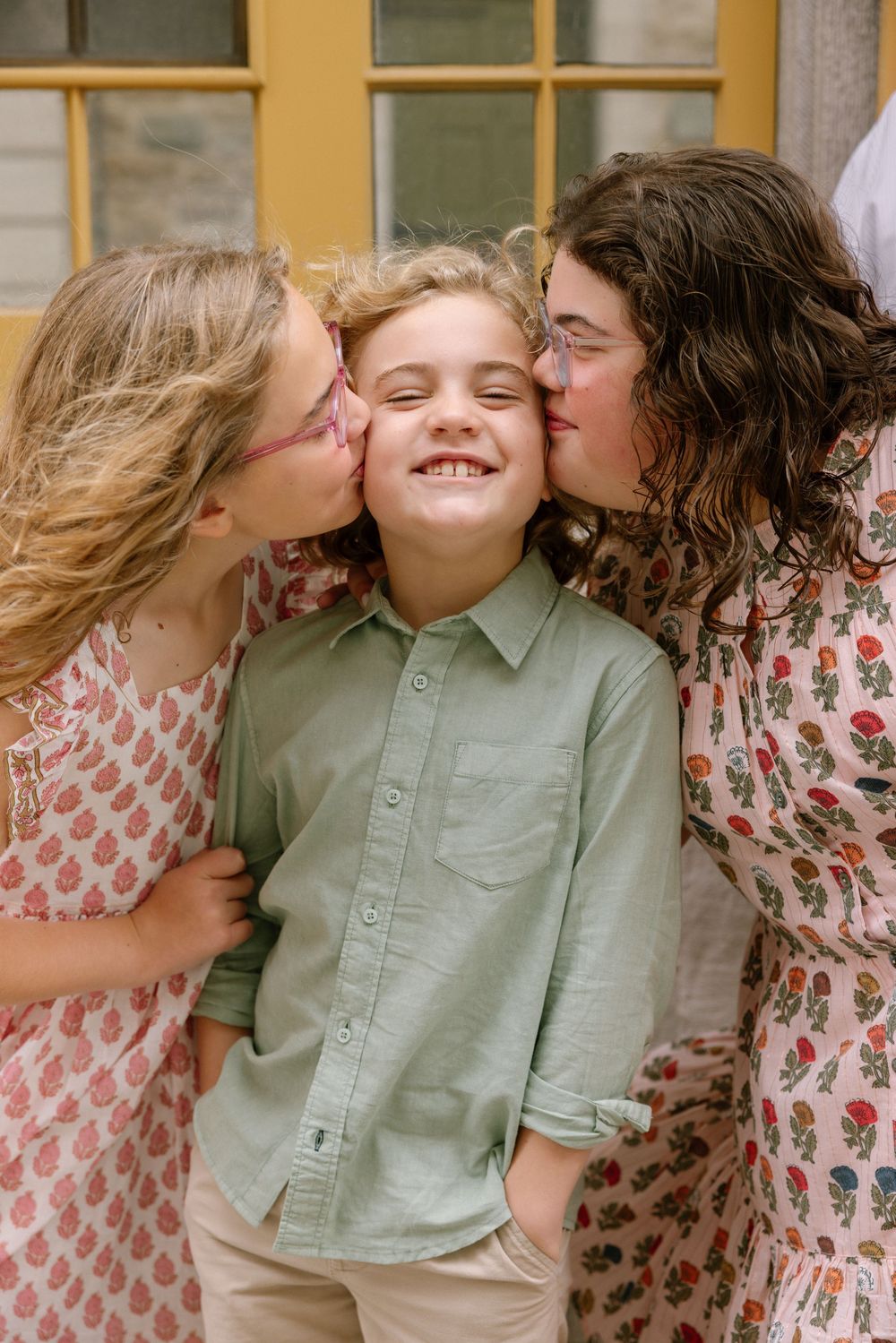 Two girls kissing a smiling boy on the cheeks outdoors, all wearing floral and pastel outfits.