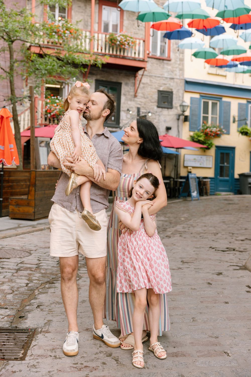 Happy family of four posing on a charming cobblestone street with colorful umbrellas overhead in Old Quebec City.