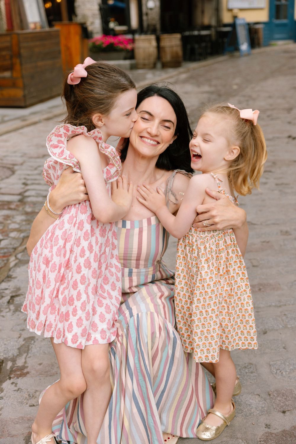A smiling mother in a striped dress hugs her two young daughters in pink dresses on a cobblestone street.
