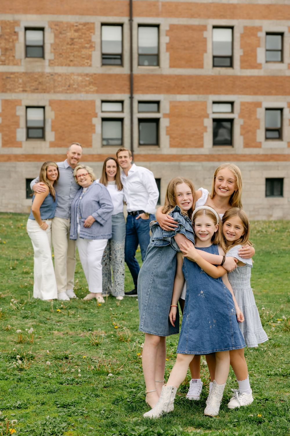 Family portrait with four girls hugging in front, grandparents and parents standing behind on green lawn near brick building.