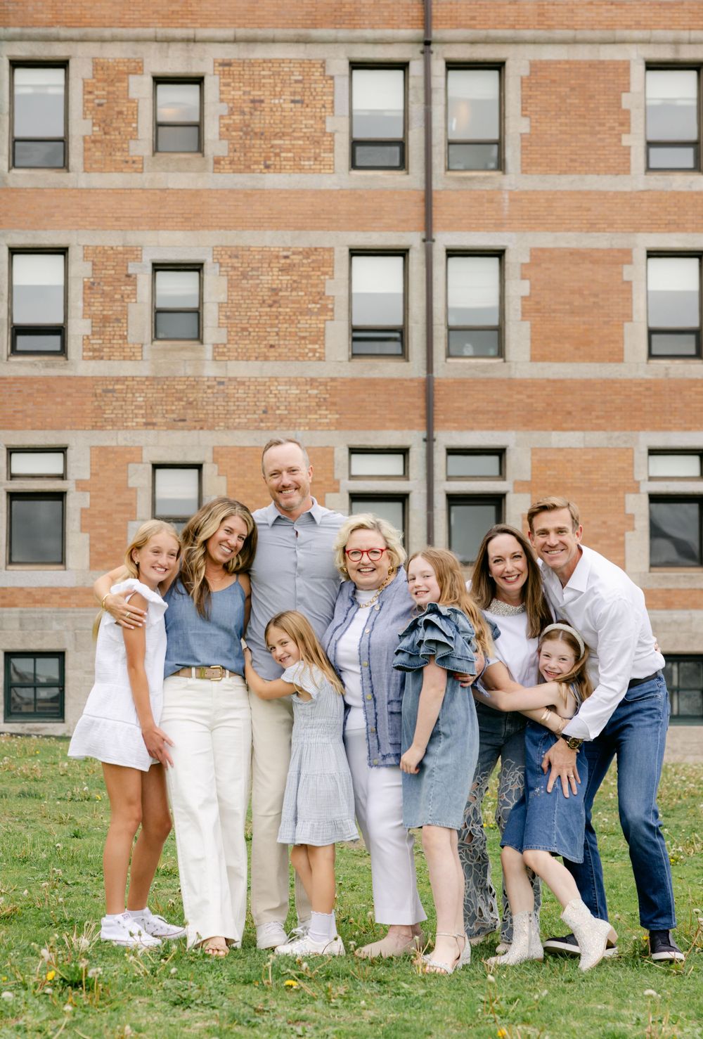 Large family group portrait smiling together outdoors in front of brick building on green grass.