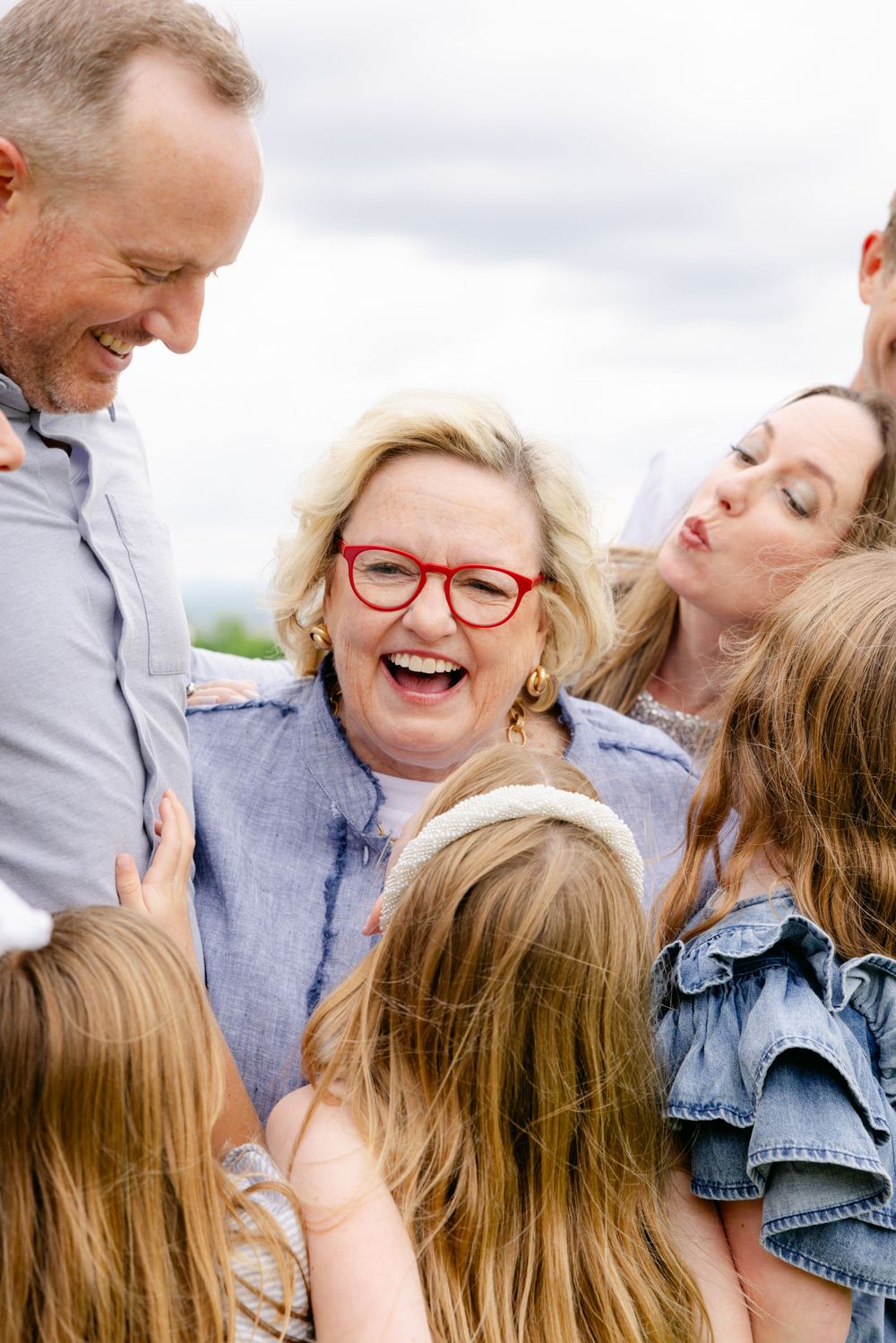 Joyful family group hug with laughing blonde woman in red glasses surrounded by children and adults outdoors.