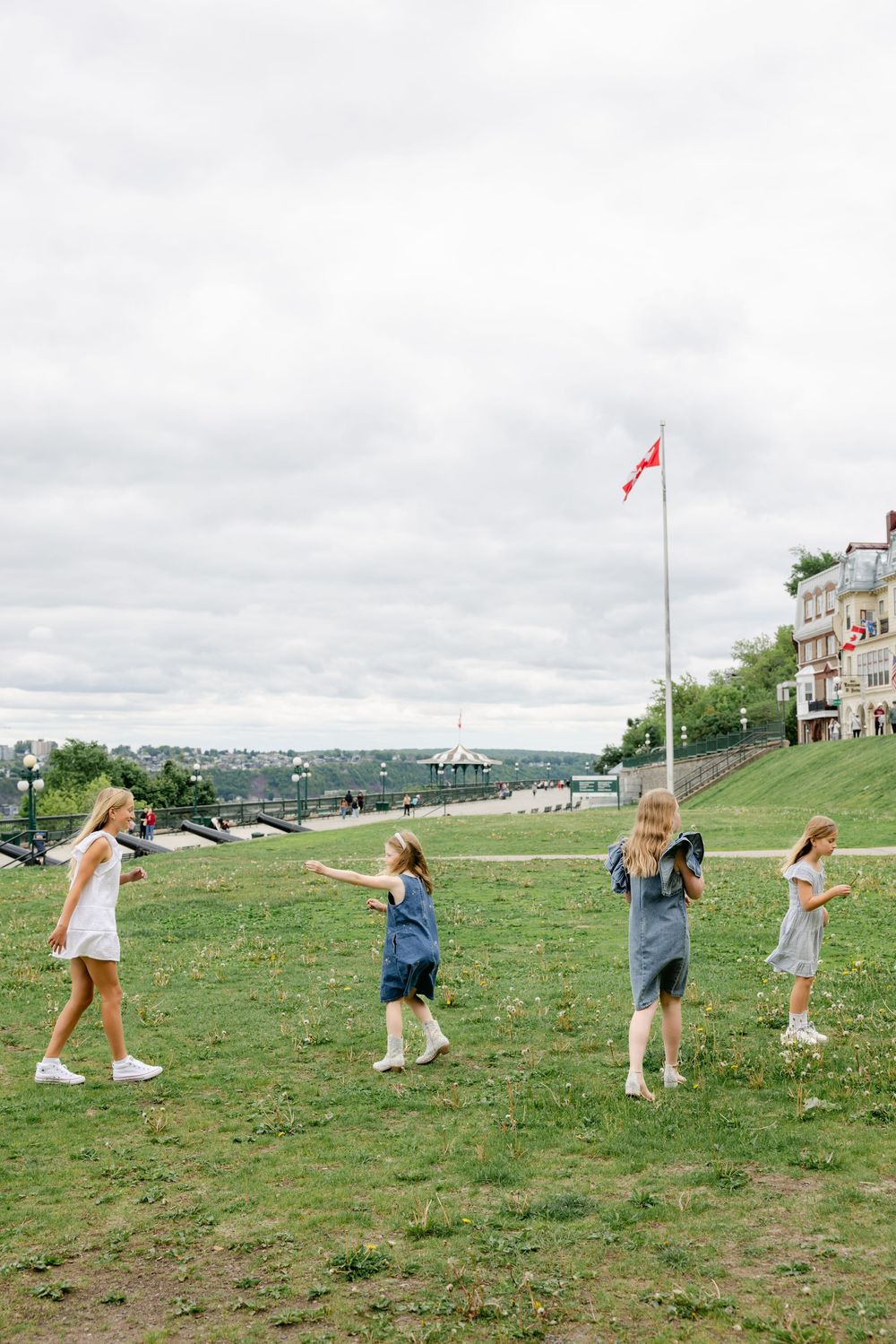 Four girls playing on a green lawn near a flagpole and waterfront promenade under a cloudy sky.