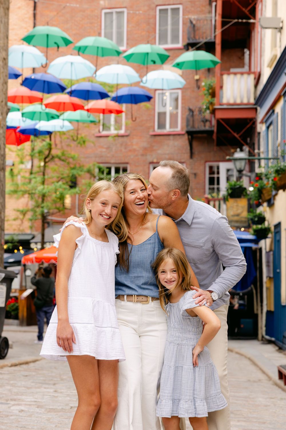 Family of four posing on cobblestone street under colorful umbrella display in charming city setting.