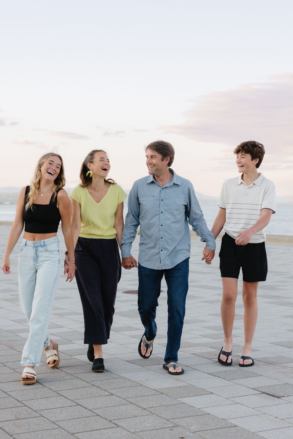 Happy family of four walking together on a seaside promenade at sunset, laughing and holding hands.