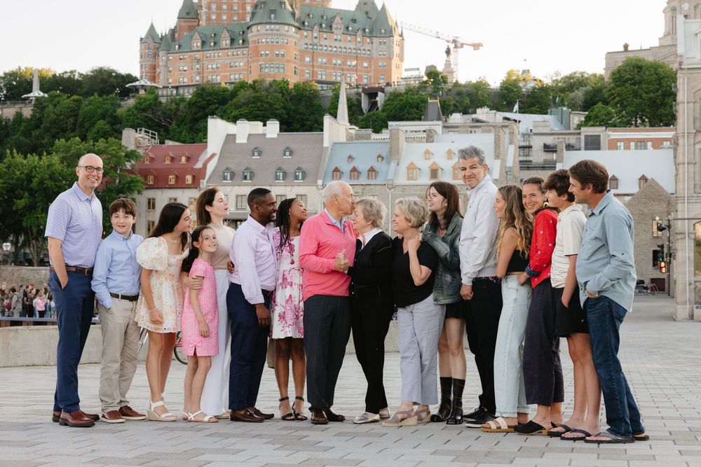 Large family group posing outdoors with Chateau Frontenac and Quebec City skyline in background on sunny day.
