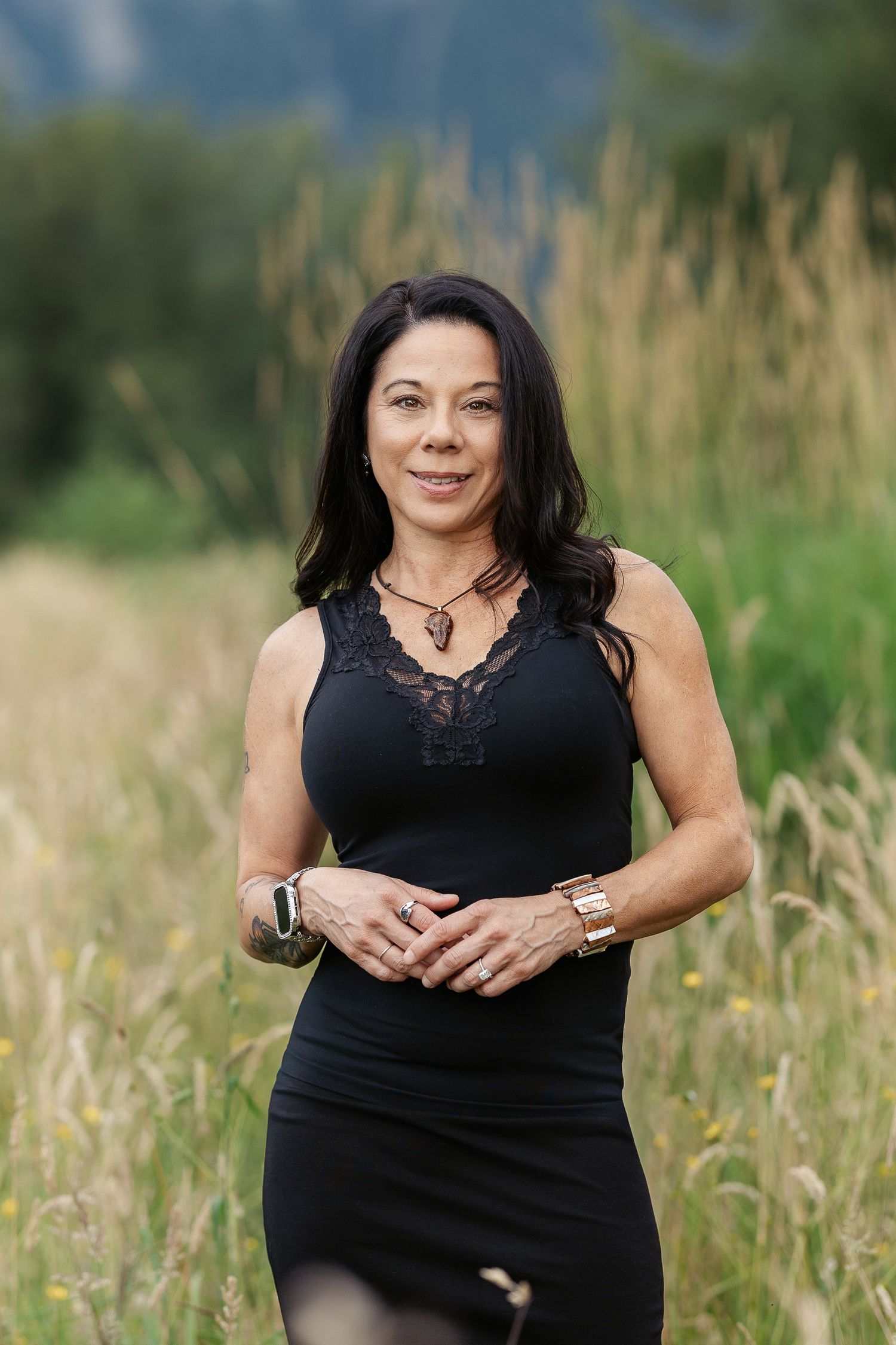 A woman in a black sleeveless dress stands in a golden field, smiling confidently with mountains in the background.