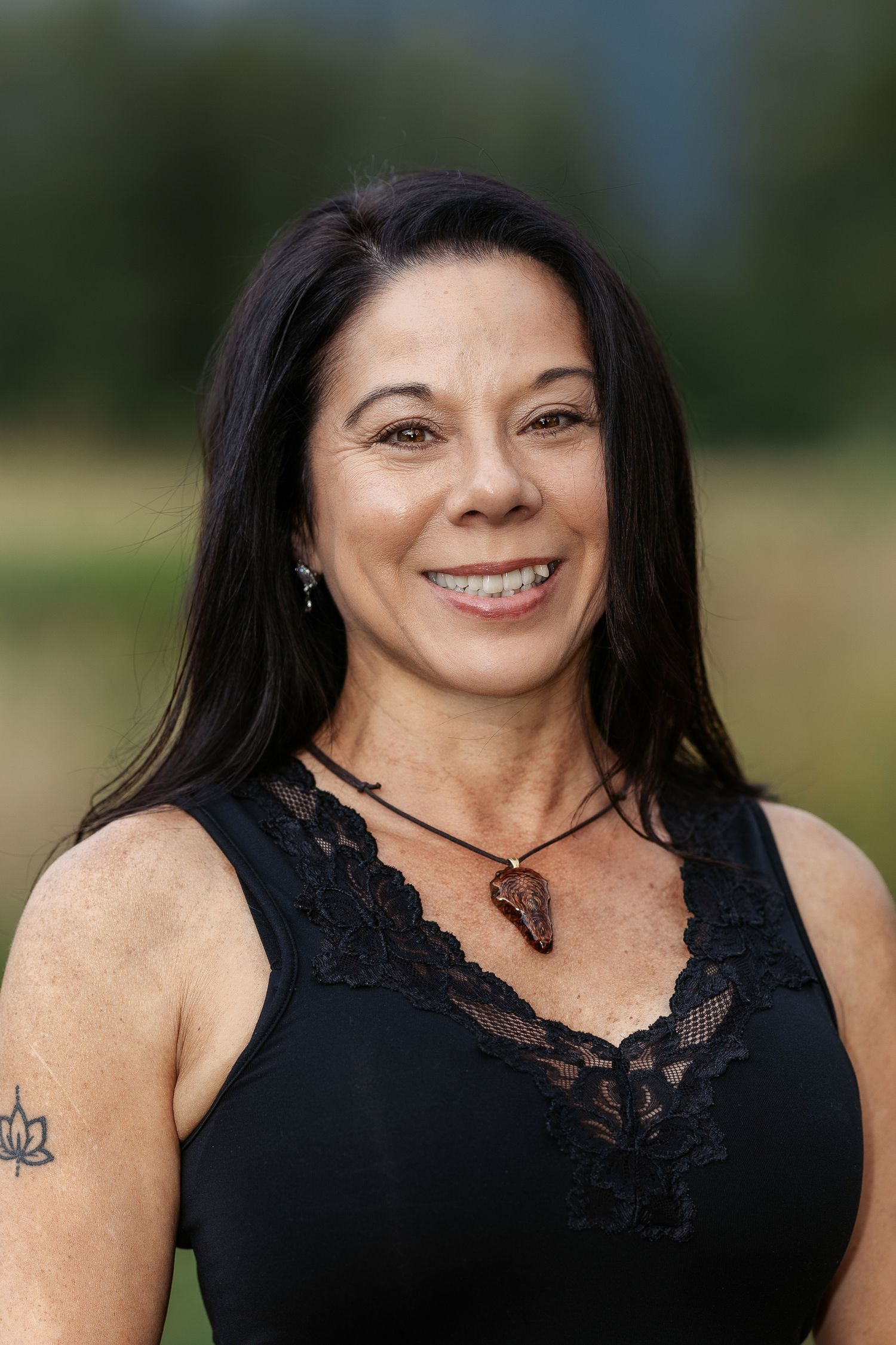 Smiling woman with dark hair wearing a black lace top and arrowhead necklace, posing outdoors with a green background.