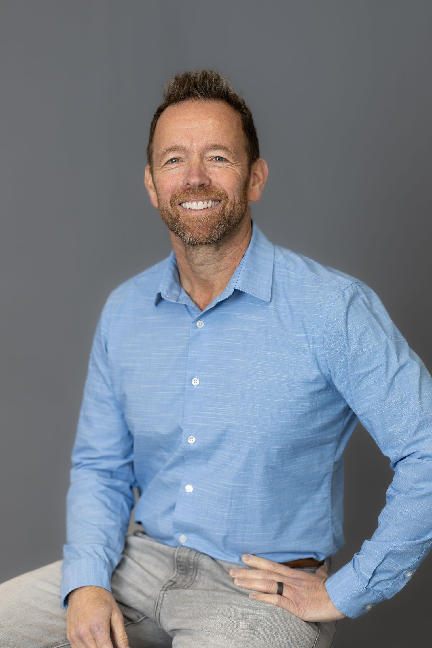 Smiling middle-aged man with beard wearing a blue button-up shirt and jeans, posed against a gray background.