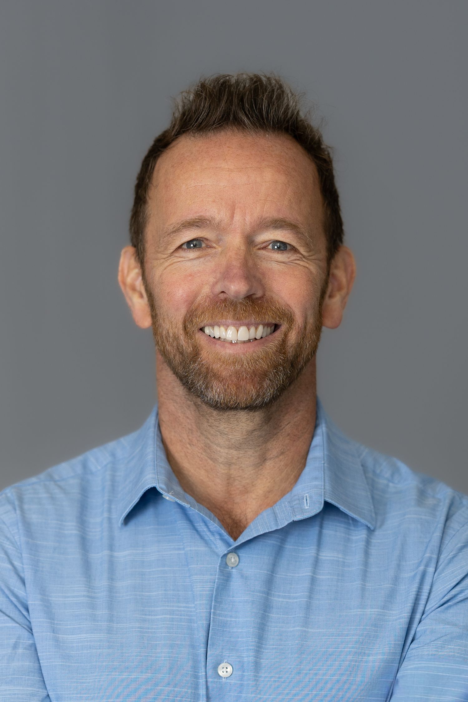 Smiling middle-aged man with beard wearing a blue button-up shirt against a gray background.