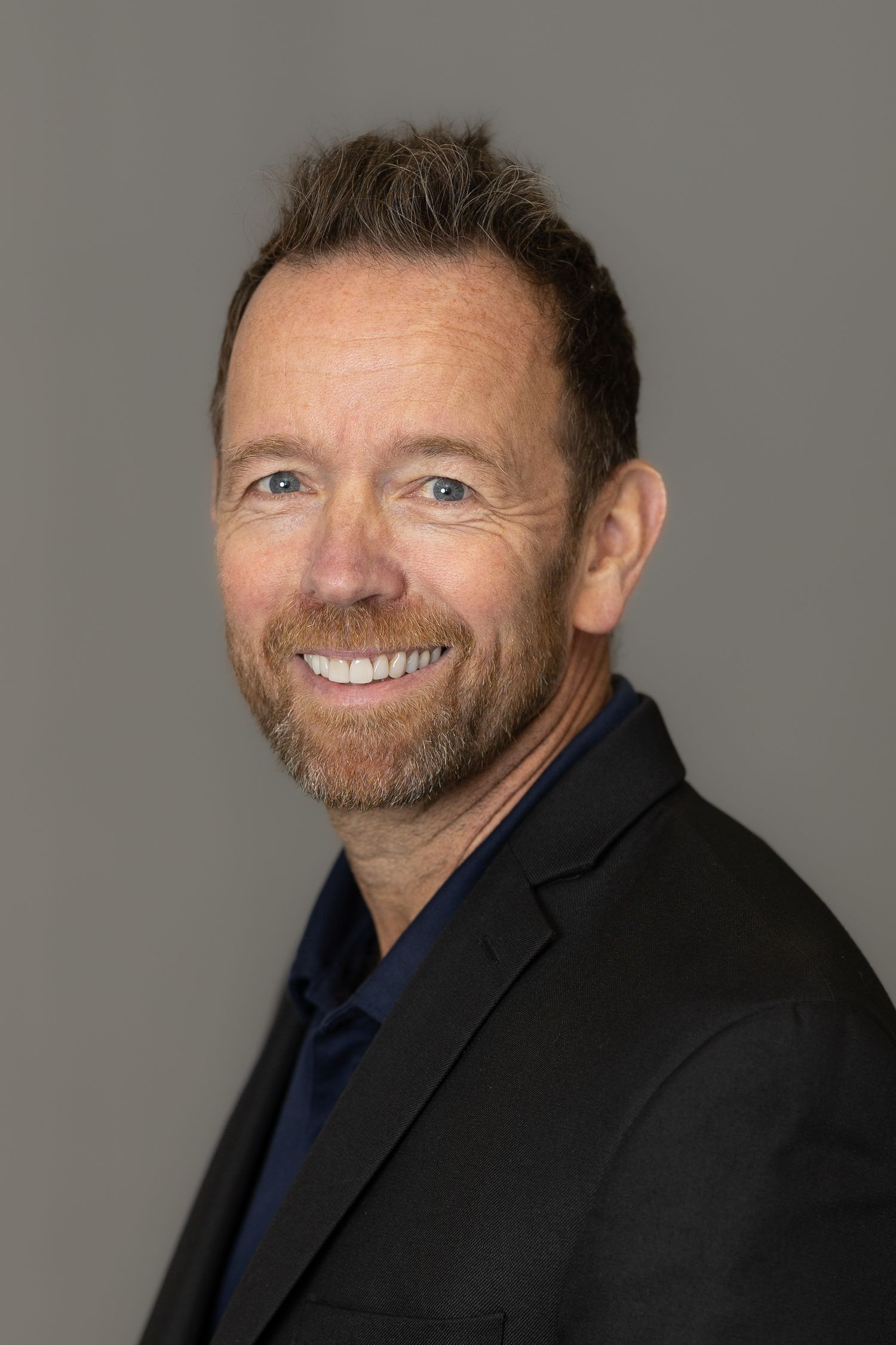 Smiling middle-aged man with beard wearing black blazer poses for professional headshot against gray background.