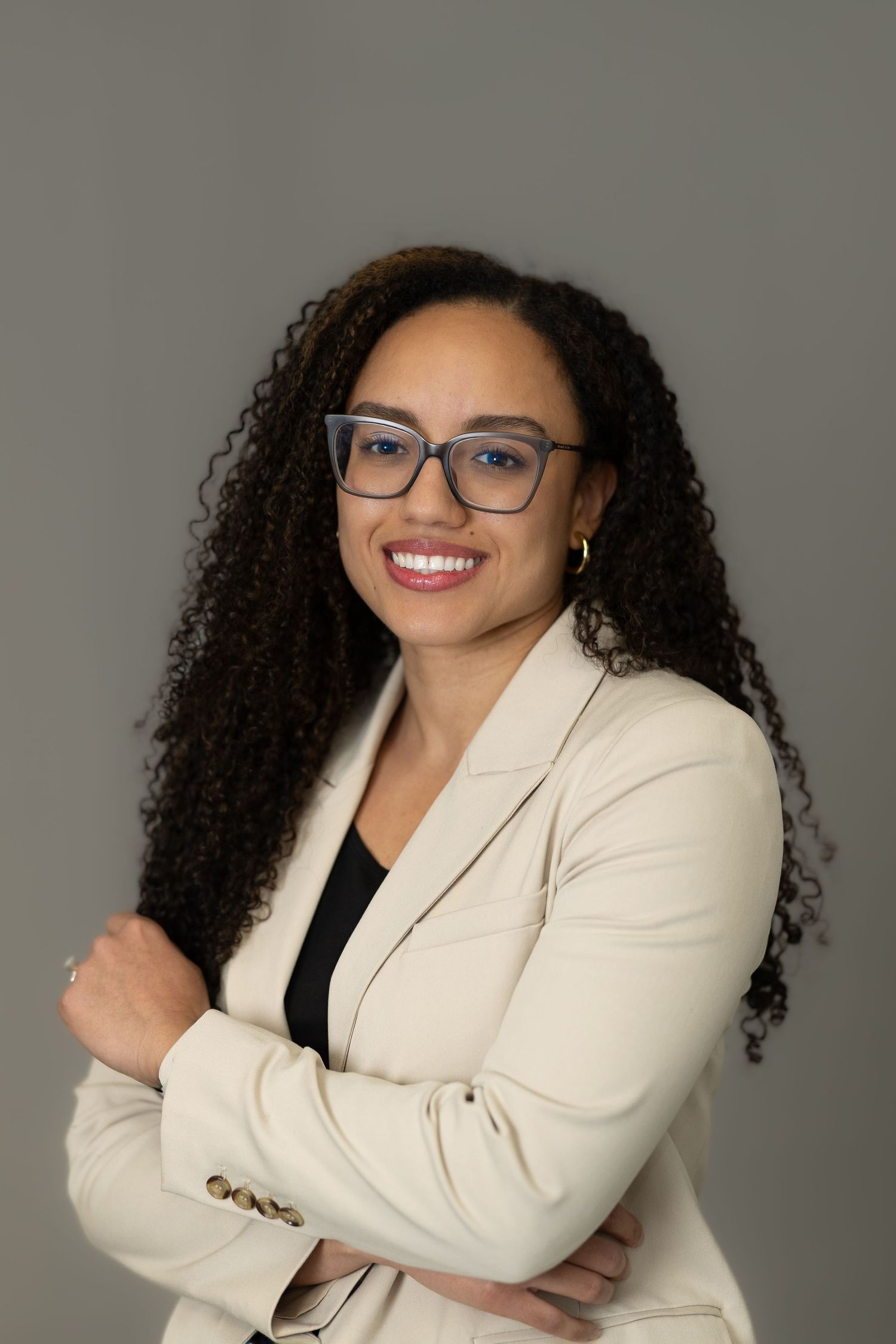Professional headshots of someone in a cream blazer and black top wearing glasses against a gray background.