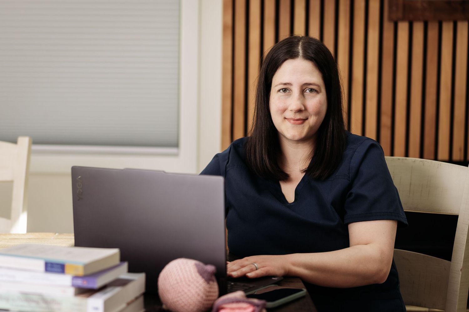 Woman in navy scrubs smiles at laptop while sitting at desk with books and pink yarn nearby.