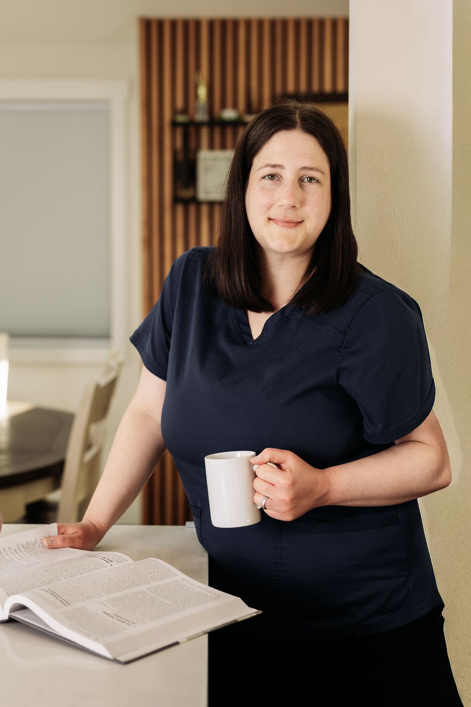 Woman in navy scrubs holding a white mug while reviewing an open book at a medical office counter.
