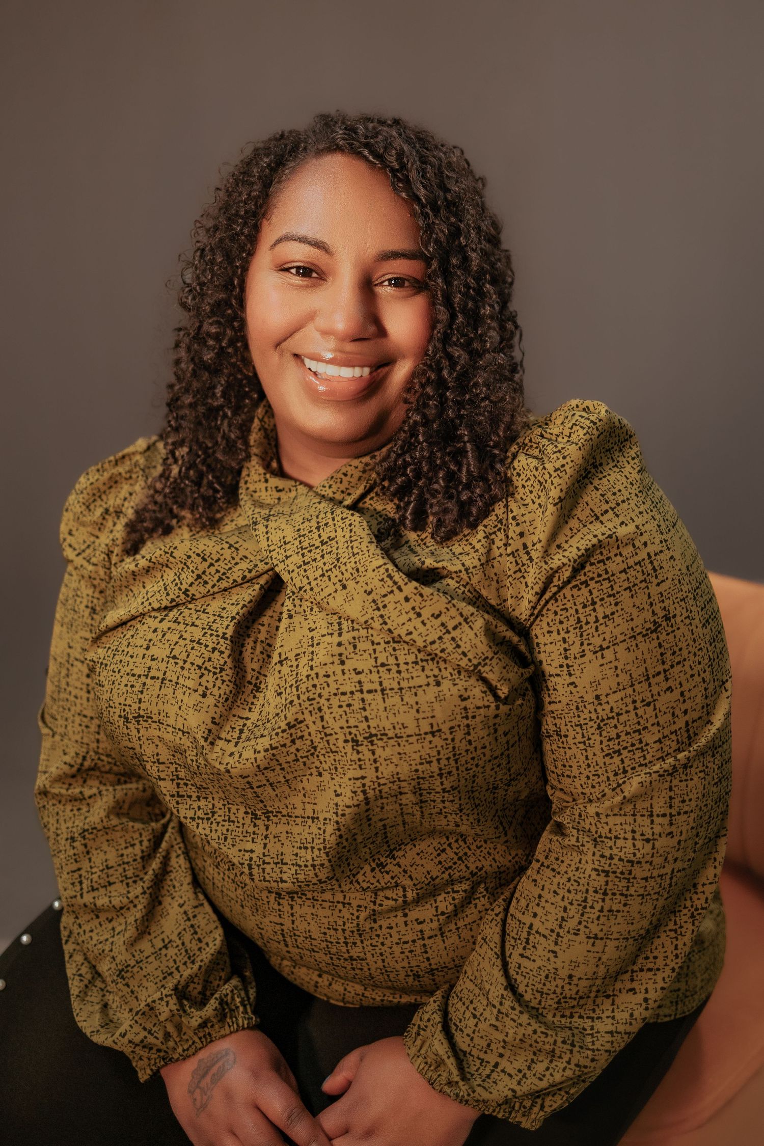 A smiling person wearing a mustard yellow textured sweater with voluminous sleeves poses against a brown backdrop.