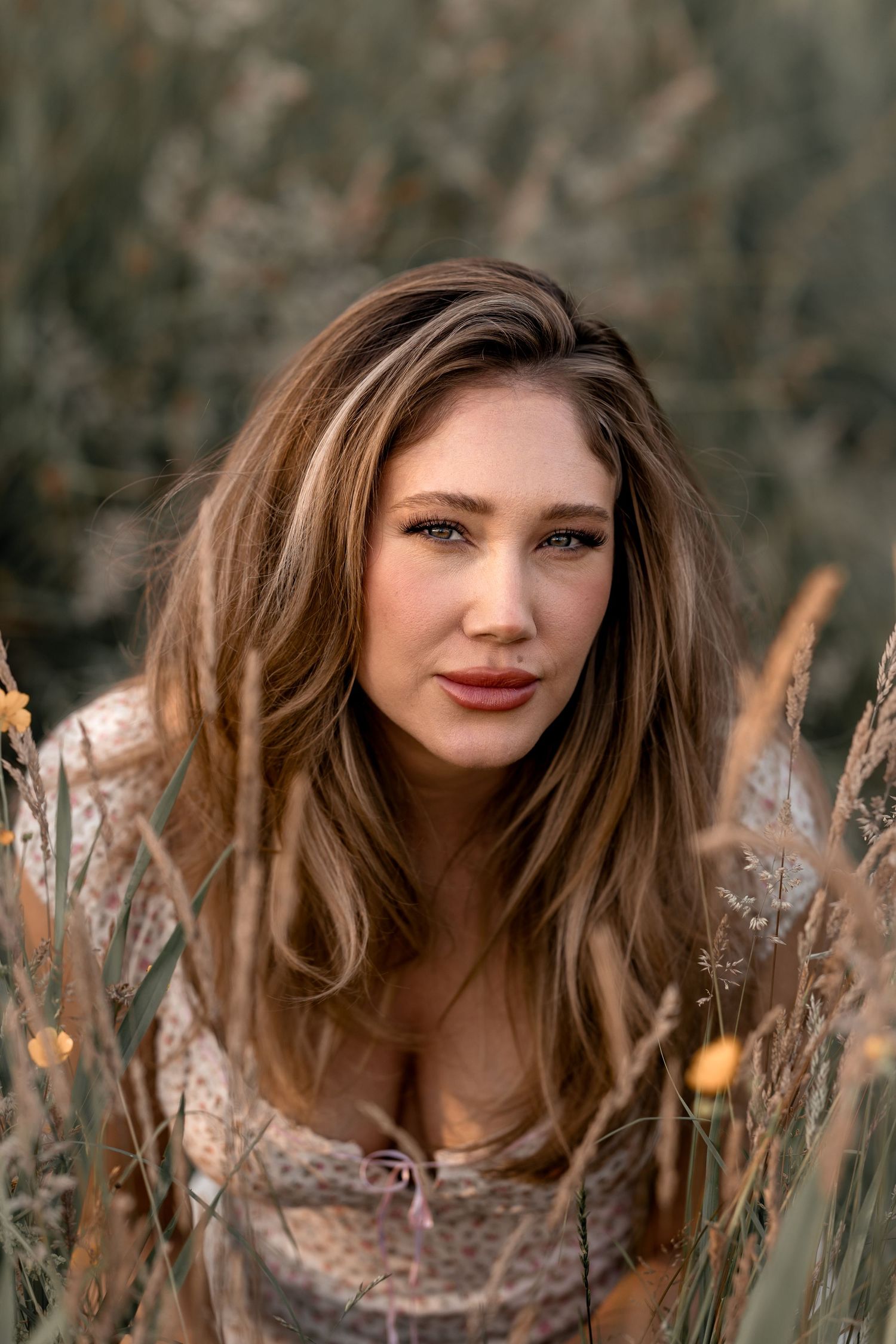 Young woman with long brown hair poses outdoors among tall wild grasses in soft natural light.