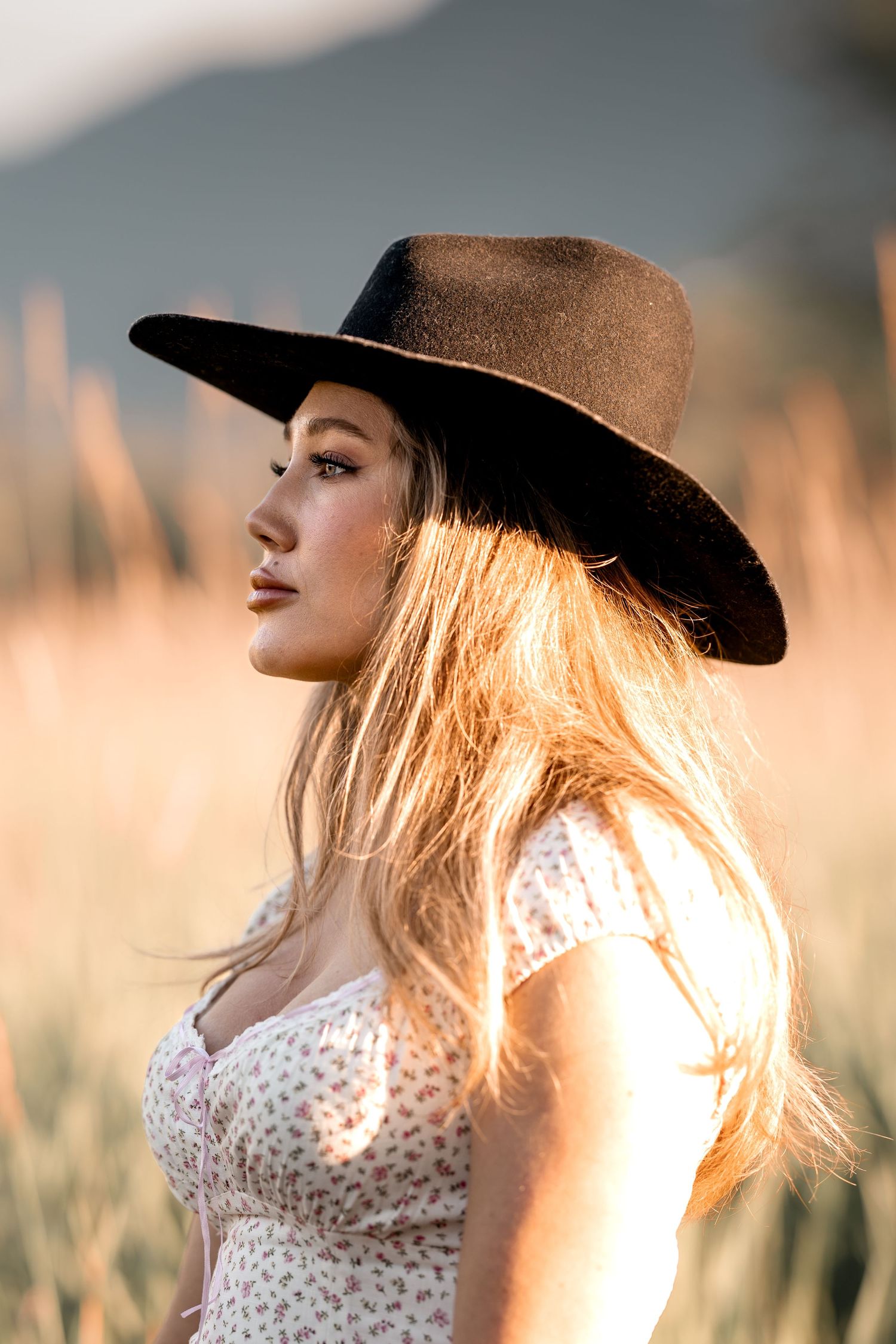 Woman in black wide-brim hat and floral dress standing in golden field at sunset.