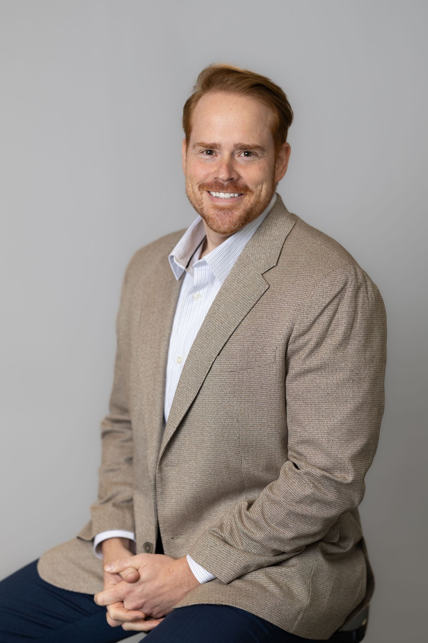 Smiling redhead man in beige blazer and white shirt sits confidently against a gray background.