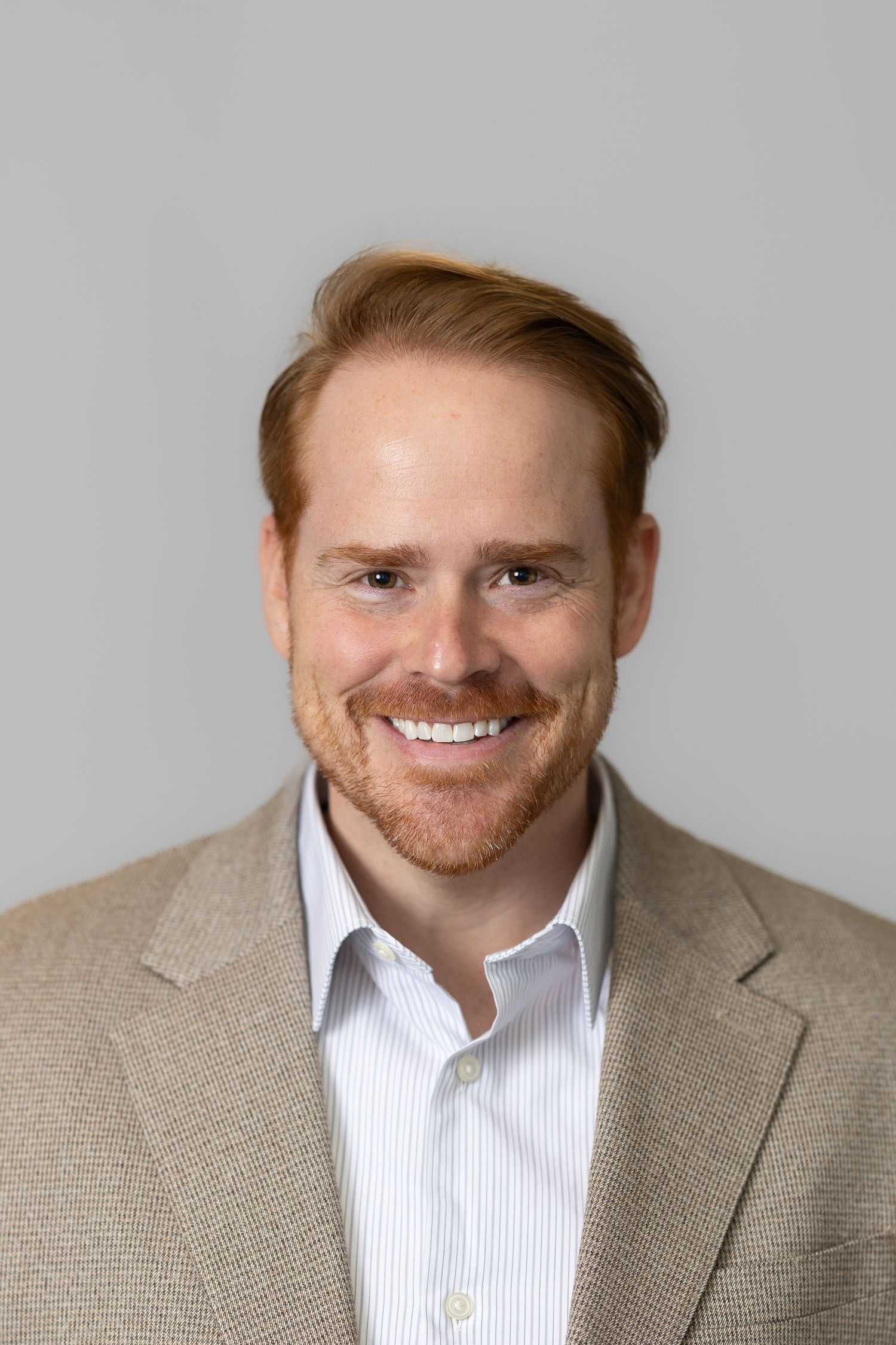 Smiling red-haired man with beard wearing a tan blazer and striped shirt against a gray background.