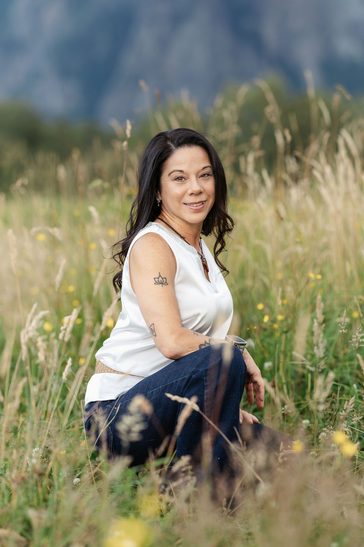 Woman with dark hair sits smiling in a wildflower meadow, wearing a white sleeveless top and jeans, with mountains behind.