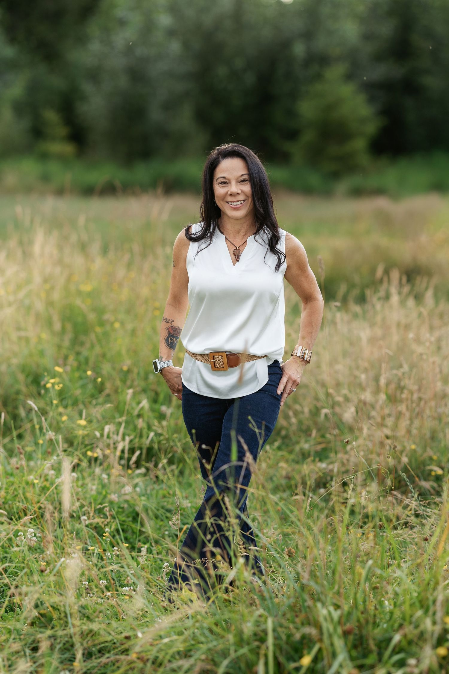 Woman in white sleeveless top and jeans with tan belt smiling in a lush green field.