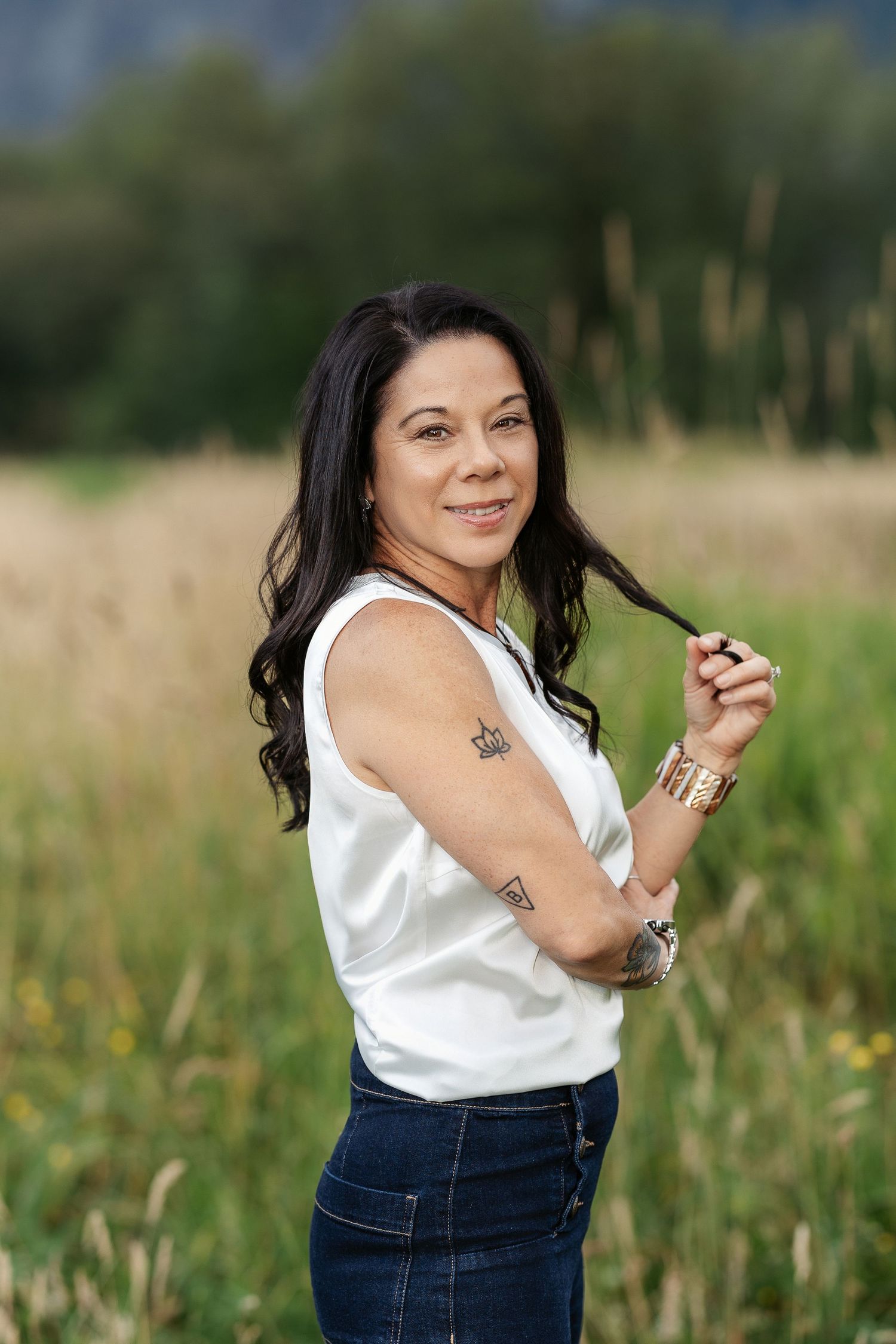 Woman in white sleeveless top and jeans poses in a field, showing tattoos and gold bracelets.