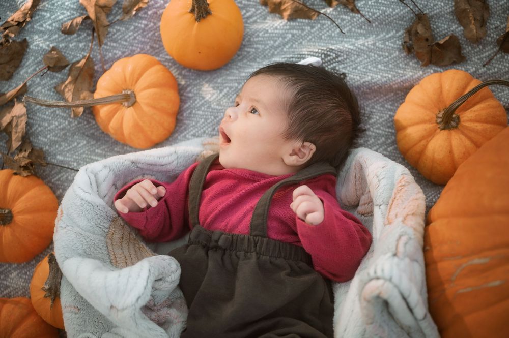 A small child lays on a white blanket surrounded by orange pumpkins at a pumpkin patch.