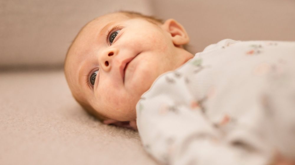 A small baby lying on beige bedding wearing a white onesie with a small smile.
