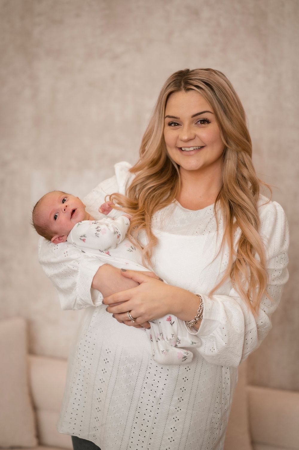 A woman in a white sweater holds a newborn baby wearing white clothing against a neutral beige background.