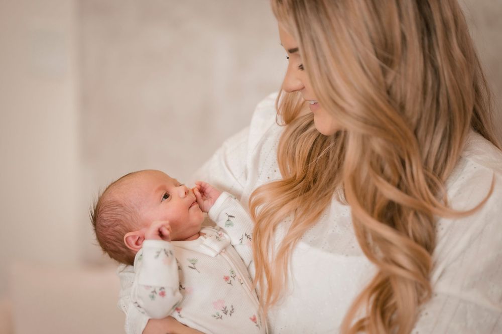 A mother in a white shirt tenderly holds her sleeping newborn baby wearing a floral onesie.