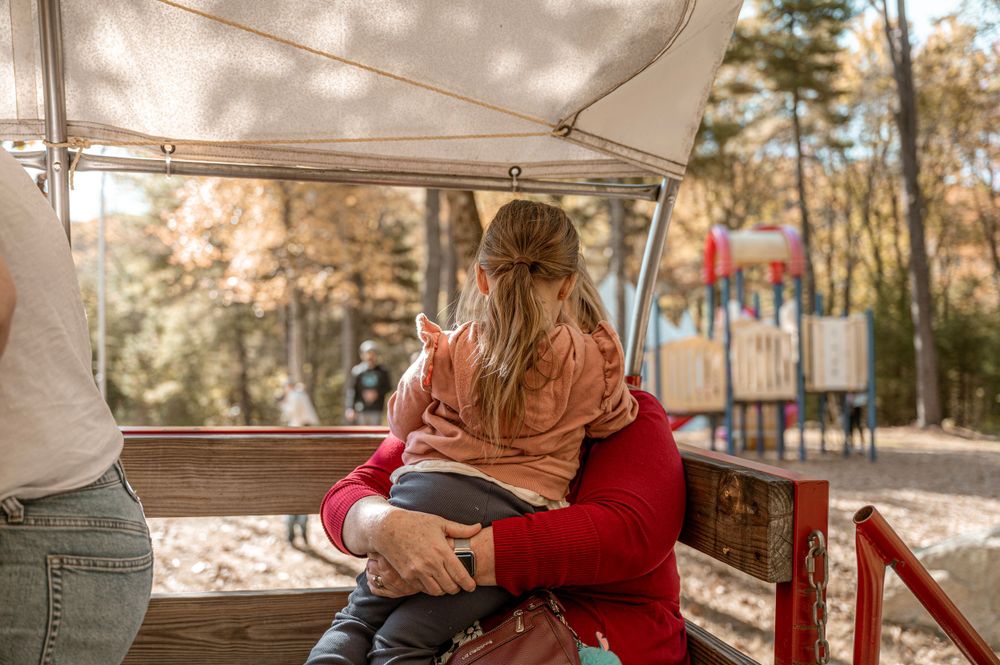 Person with long blonde hair sitting on a red bench inside a covered outdoor seating area on a sunny day.