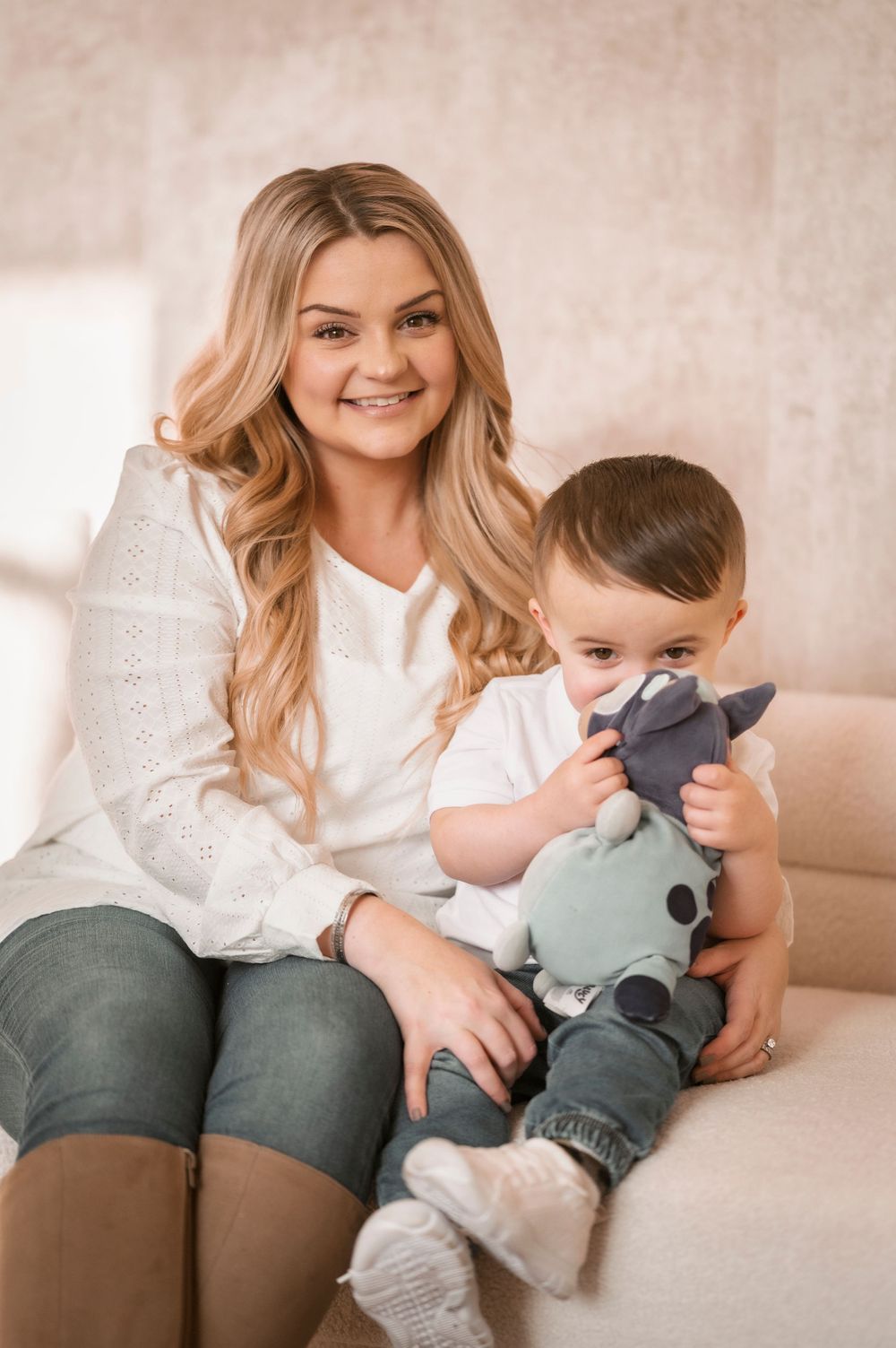 A parent and young toddler sit together on a couch, the child holding a plush toy while wearing casual clothing.
