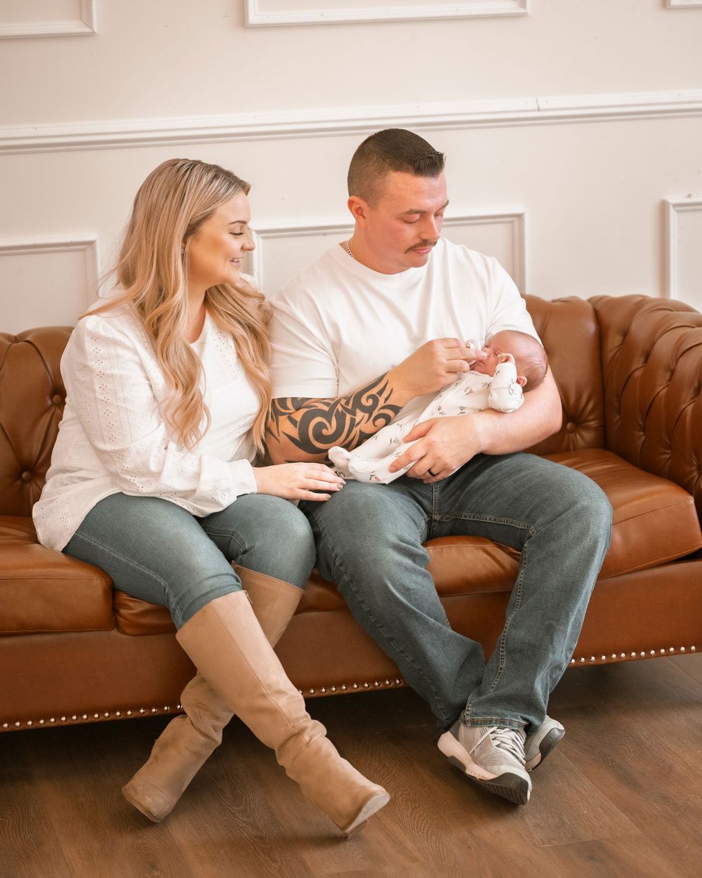 A couple sits on a brown leather couch holding a newborn baby in a cozy living room setting.