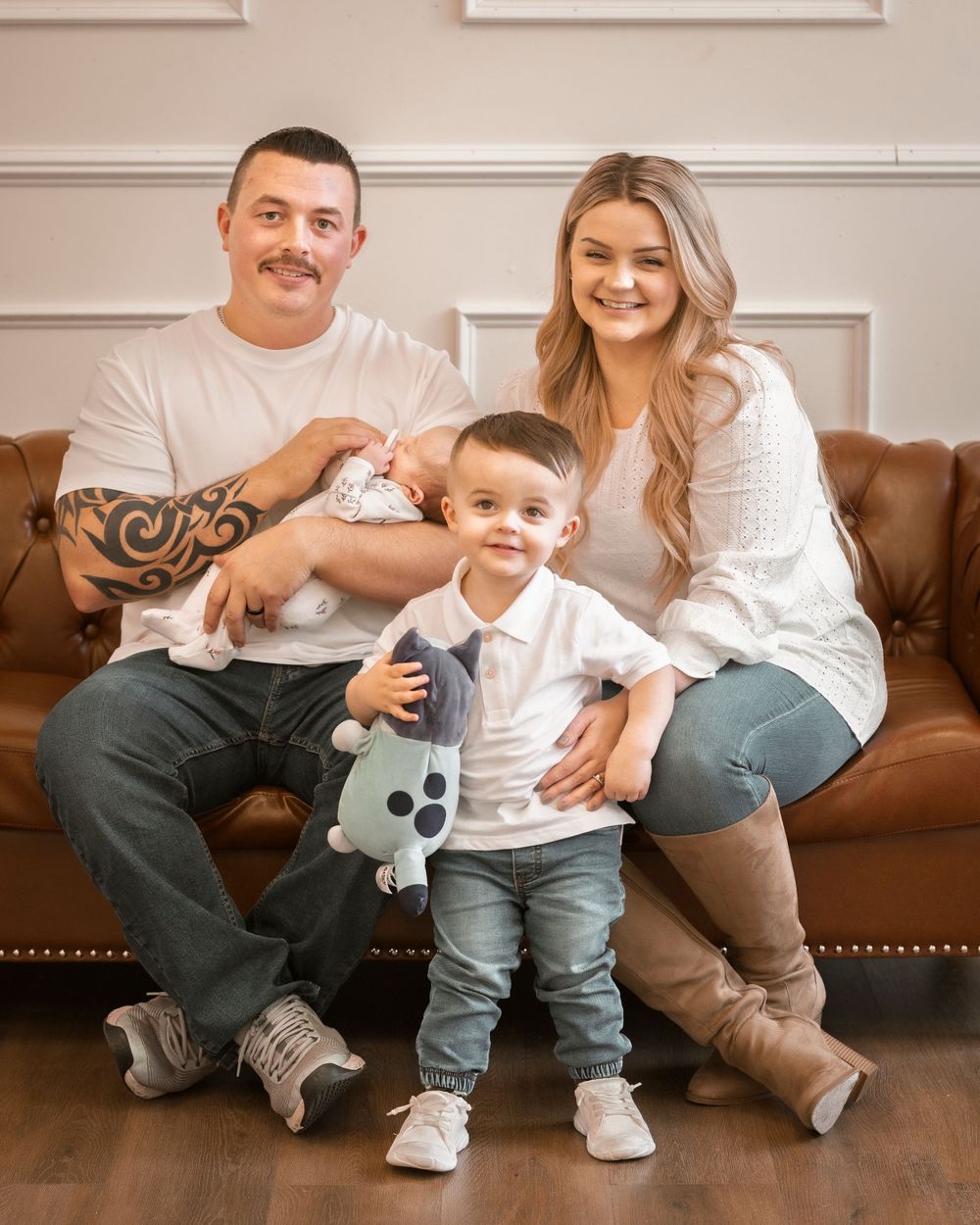 A young family sits together on a brown leather couch, wearing casual attire and smiling warmly at the camera.