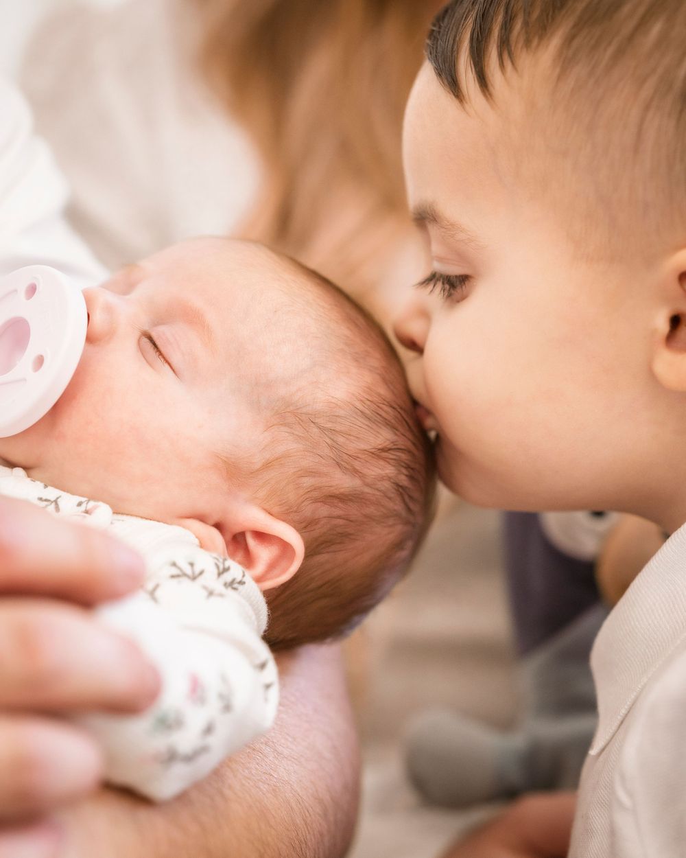 A young child gives a gentle kiss to a sleeping baby with a pacifier.