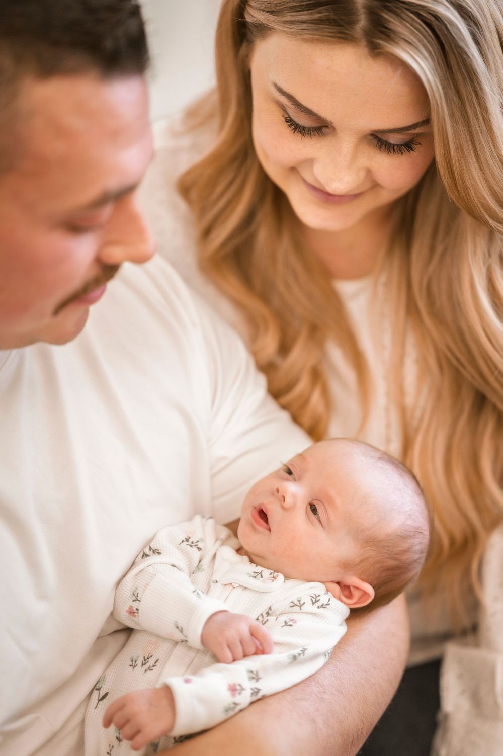 A couple gently holds their newborn baby dressed in a patterned onesie while sharing an intimate moment together.