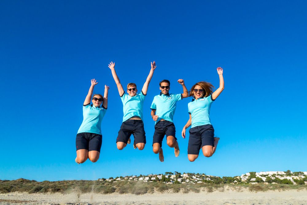 Four people in matching blue shirts and shorts jumping joyfully together against bright blue sky at beach.