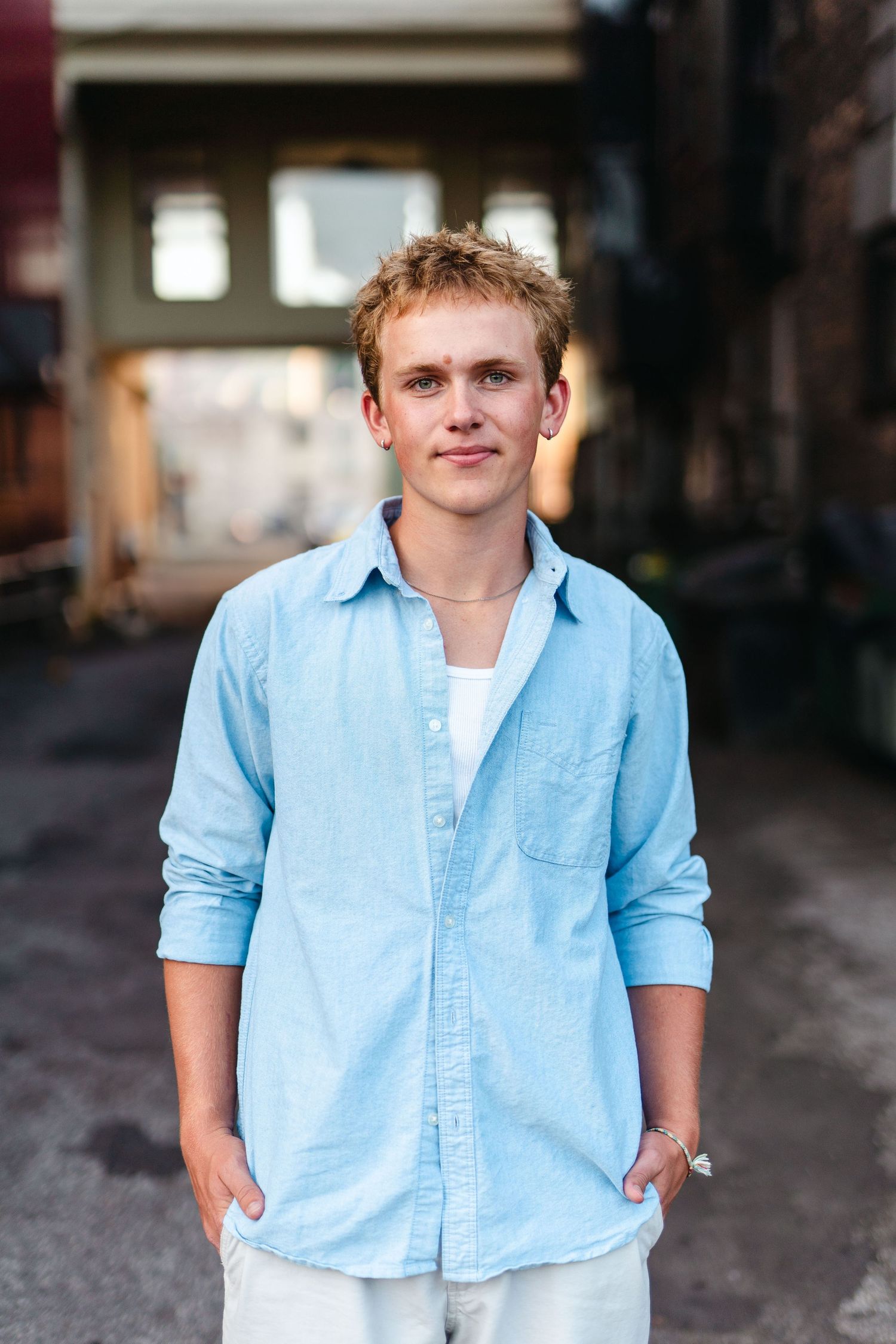 A person in a light blue button-down shirt and white pants stands in a casual pose against an industrial background.