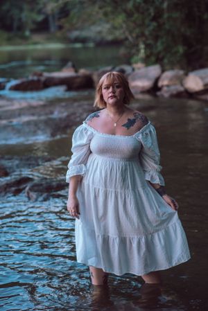 Woman in white dress standing in a flowing stream surrounded by rocks and trees.