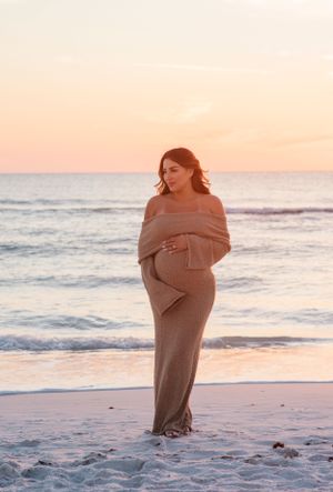 Full-length maternity portrait of a woman in a beige dress on a beach at sunset.