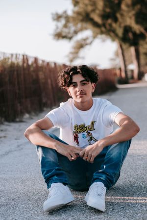 Young person in white graphic t-shirt and jeans sitting casually on pavement at sunset.